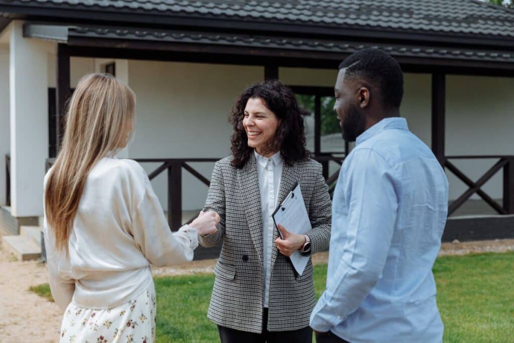 A beaming real estate agent shakes hands with a couple in front of a house